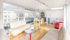 Een ruime woonkamer met een bank, een glazen salontafel en een eethoek met stoelen en een tafel. Grote ramen bieden uitzicht op gebouwen en een balkon.