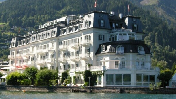 Een groot, meerhoog hotel met balkons en uitzicht op het water, omgeven door groen en heuvels.