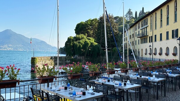 An outdoor dining area with tables and chairs, overlooking a body of water and a mountain in the background.