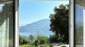 A view through a window of a lake with mountains in the background, a wooden bench, and a paved path.