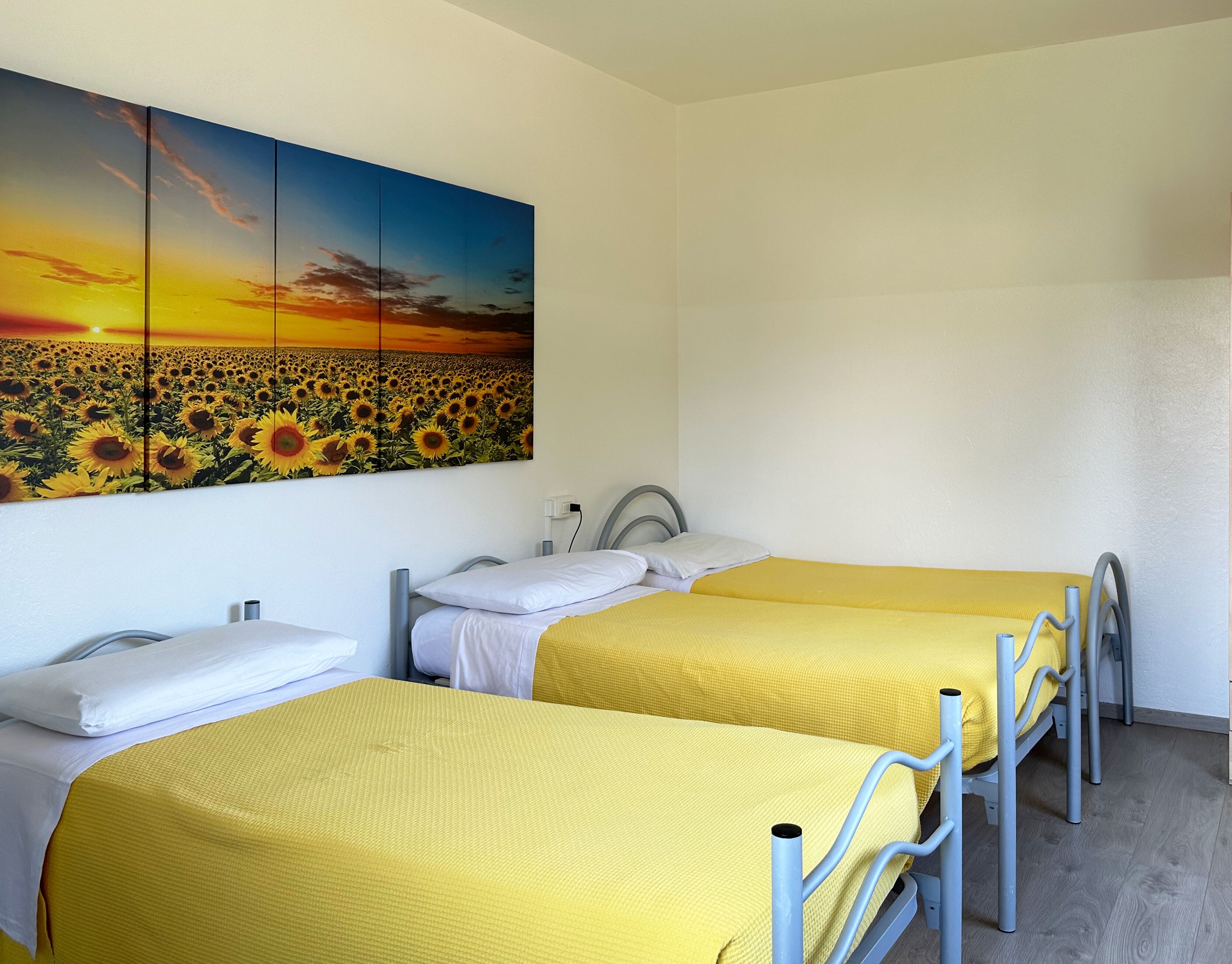 Una habitación con dos camas, cada una con cubrecama amarillo, un cuadro grande de un campo de girasoles y una pared blanca.