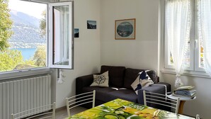 A room with a dining table covered with a fruit-themed tablecloth, a sofa with cushions, a radiator, a window with a view of a lake and mountains, and a framed picture on the wall.