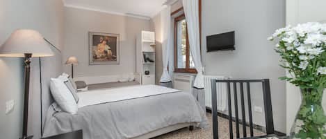 A bedroom with a bed, bedside tables, a desk, a chair, a TV, a radiator, and a window with curtains.