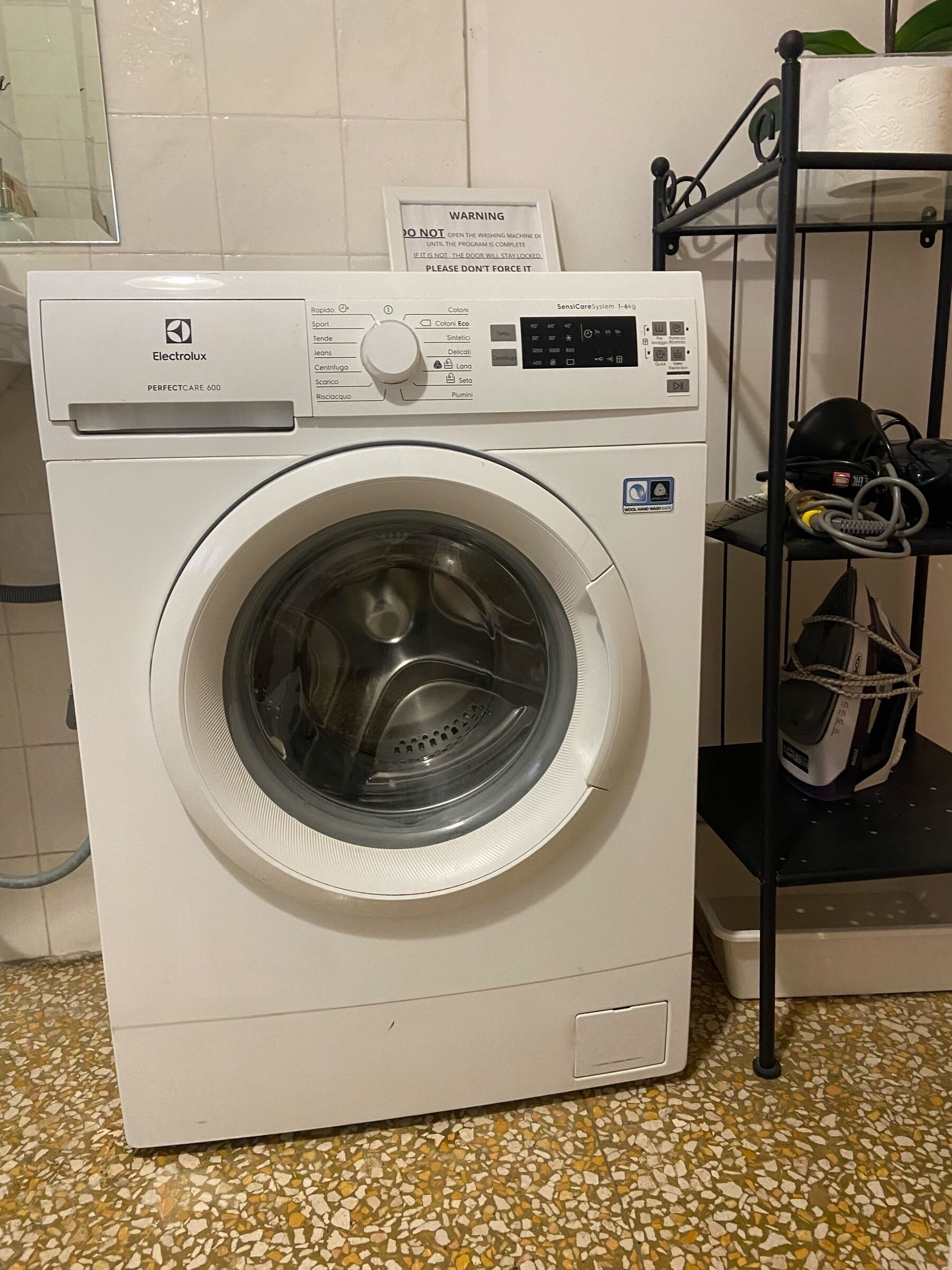 A front-loading washing machine with a control panel and a warning sign above it.