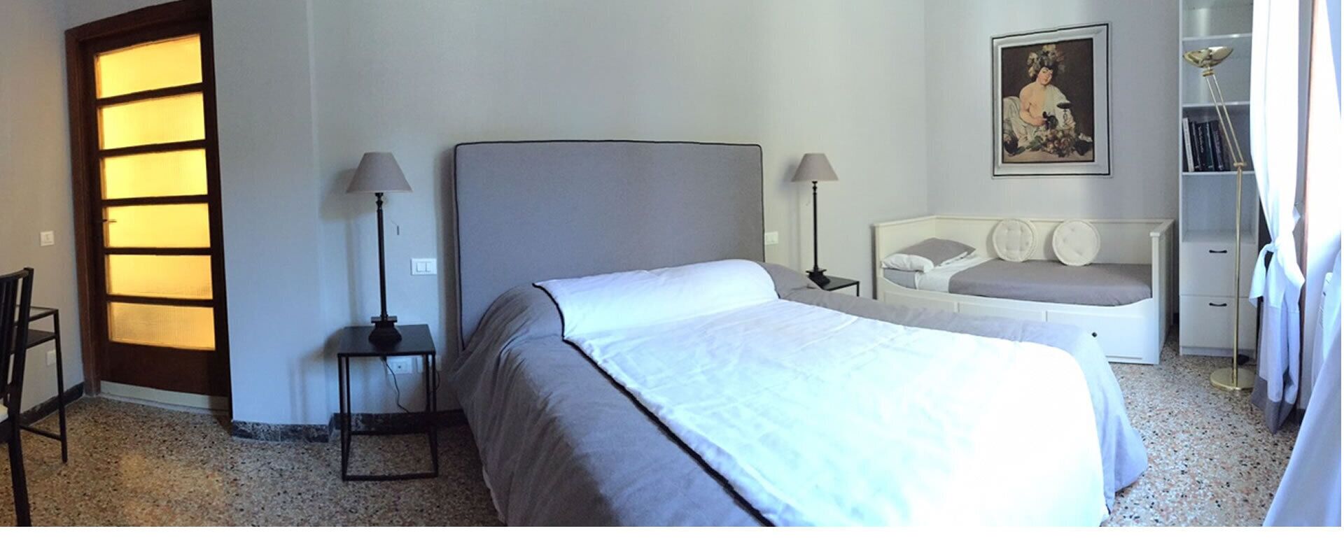 A hotel room with a bed, bedside tables, a nightstand, a chair, a desk, a lamp, a bookshelf, and a painting on the wall.