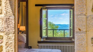 A room with a bed, a nightstand with a lamp, a window with a view of greenery and hills, and a radiator. - Montebendico 6, Emma Villas (Montegiovi - Castel del Piano (Grosseto))