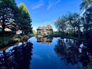 A stone building with a sloped roof, surrounded by trees and a calm water body reflecting the structure.