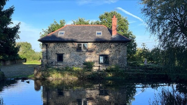 A stone house with a red roof, reflecting in a calm body of water.