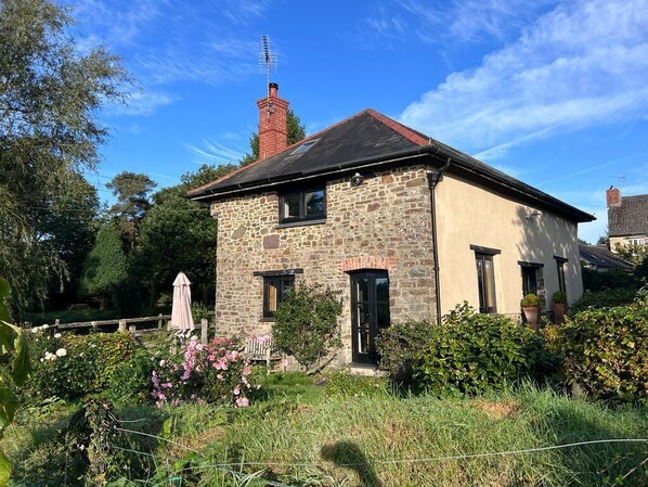 A stone house with a red chimney, surrounded by a garden with various plants and flowers.