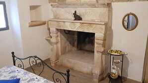 Una habitación con chimenea, una cama con cubrecama floral, una mesita con una jarrón y un espejo en la pared.