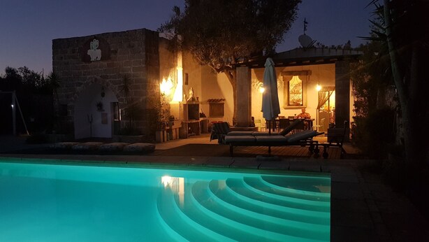 Zona junto a la piscina con un edificio de piedra, mobiliario exterior e interiores bien iluminados.