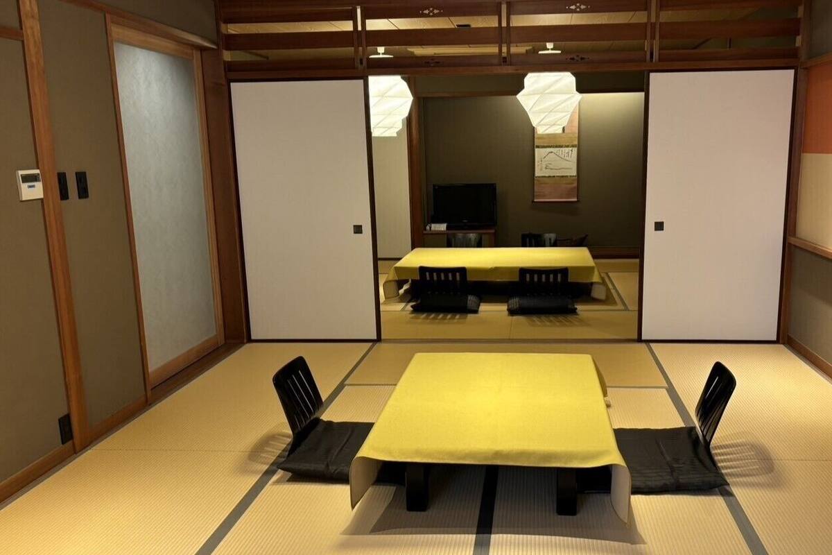 A traditional Japanese room with tatami flooring, a low table with a yellow tablecloth, black chairs, and sliding doors.