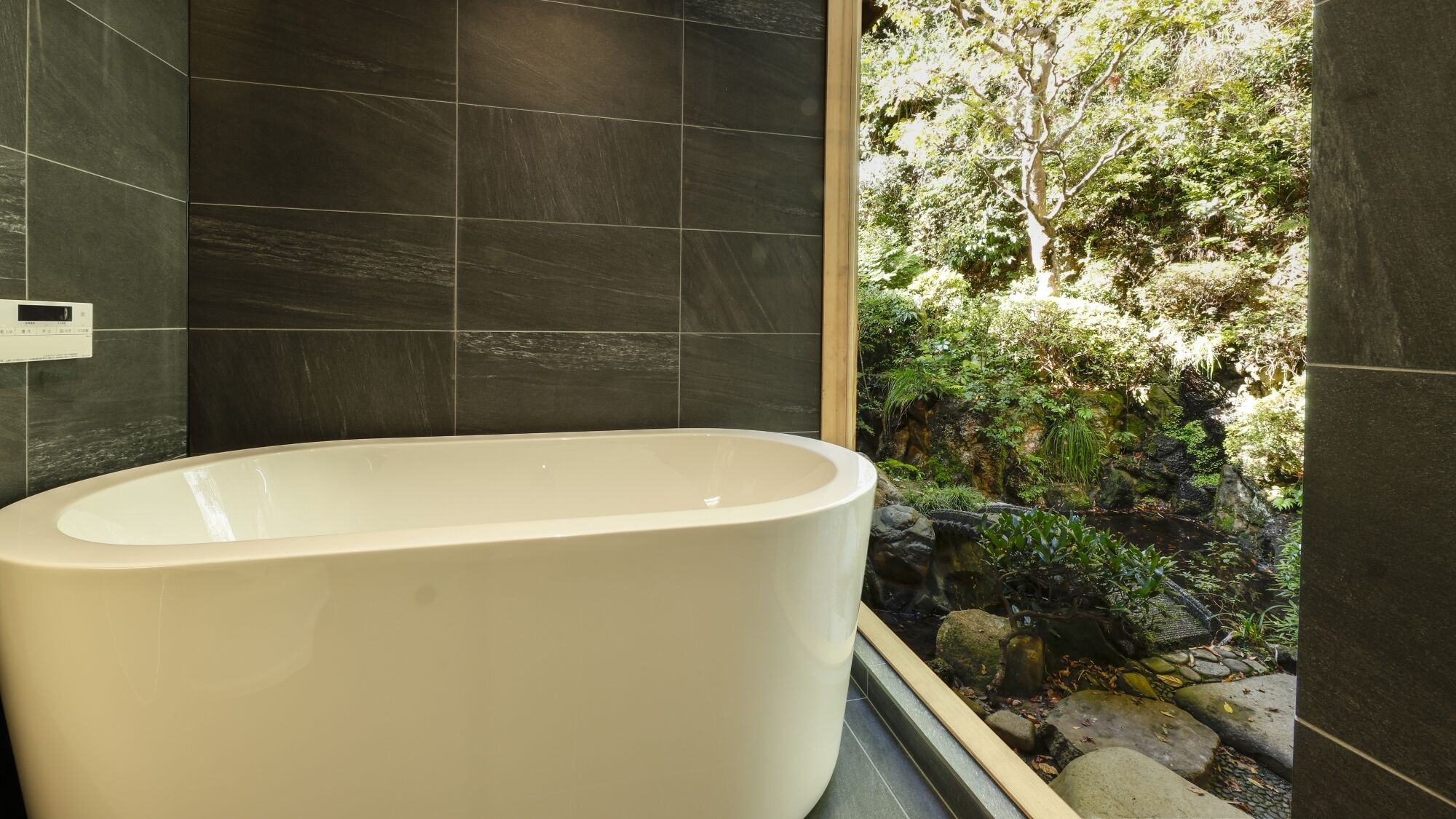 A modern bathroom with dark tiles, a freestanding bathtub, and a view of a natural outdoor area.