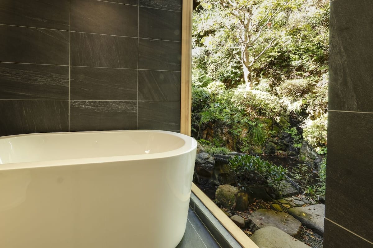A modern bathroom with dark tiles, a freestanding bathtub, and a view of a natural outdoor area.