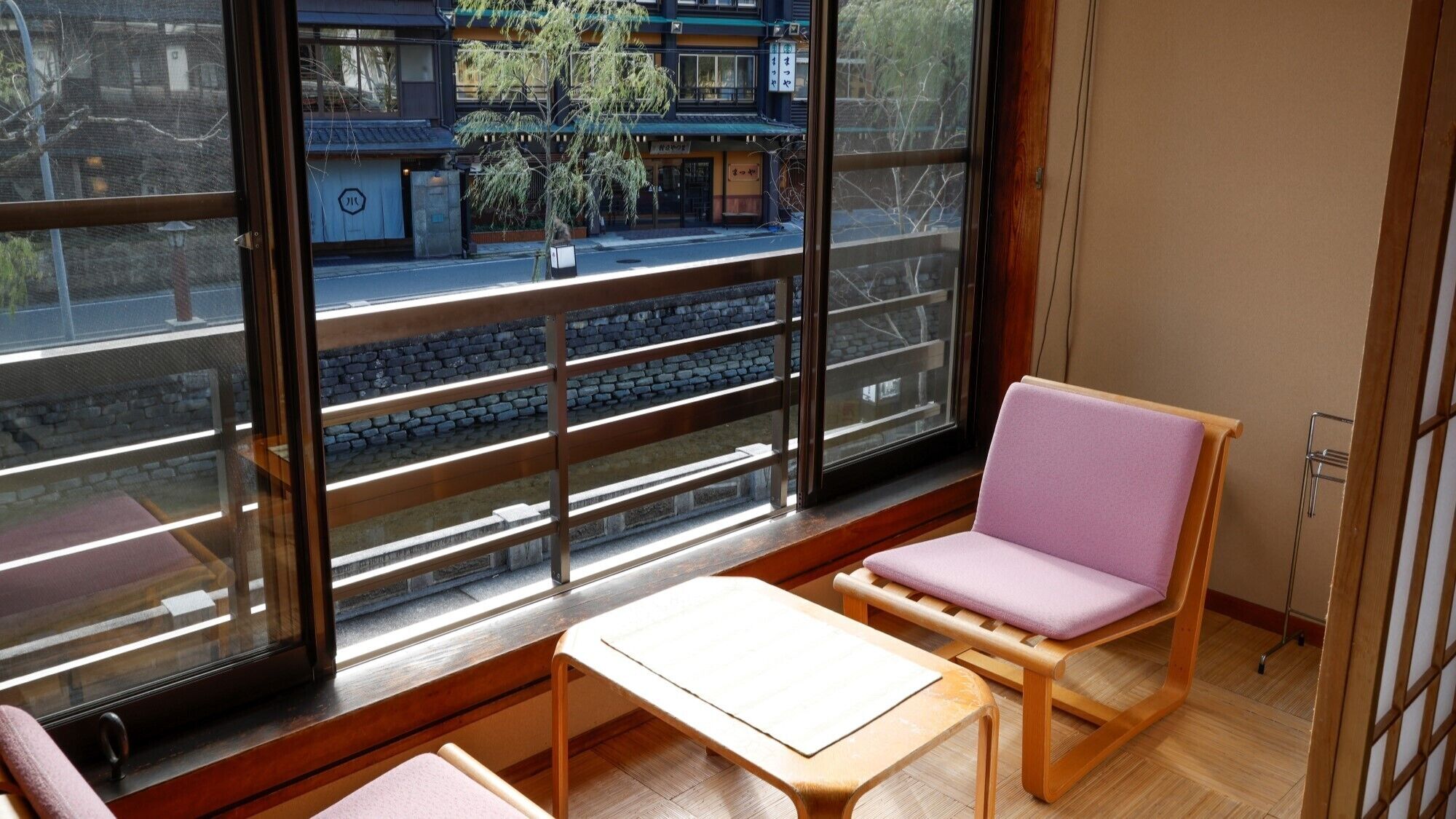 Een kamer met een groot raam, een houten bankje, een kleine tafel en een stoel met een kussen.