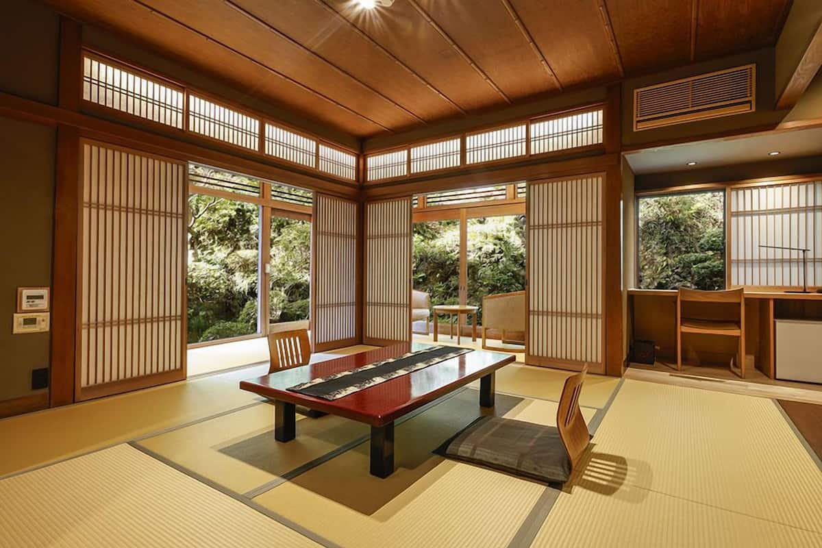 A traditional Japanese room with tatami flooring, a low table, sliding doors, and a view of greenery outside.