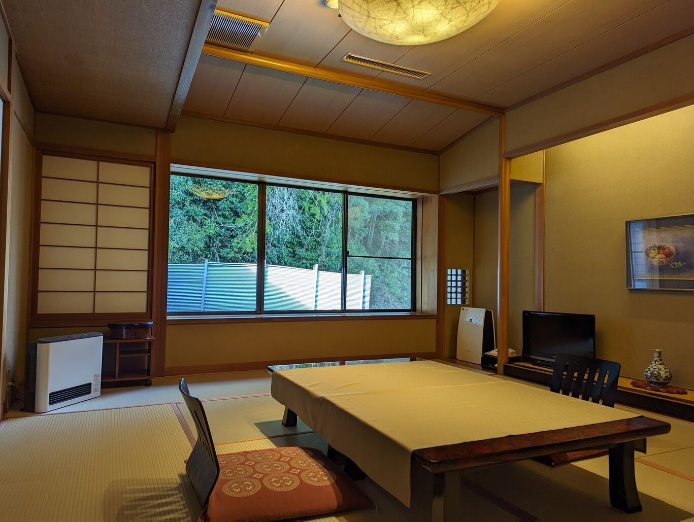 Een kamer met tatami matten, een tatami tafel, een televisie en een groot raam met uitzicht op bomen.
