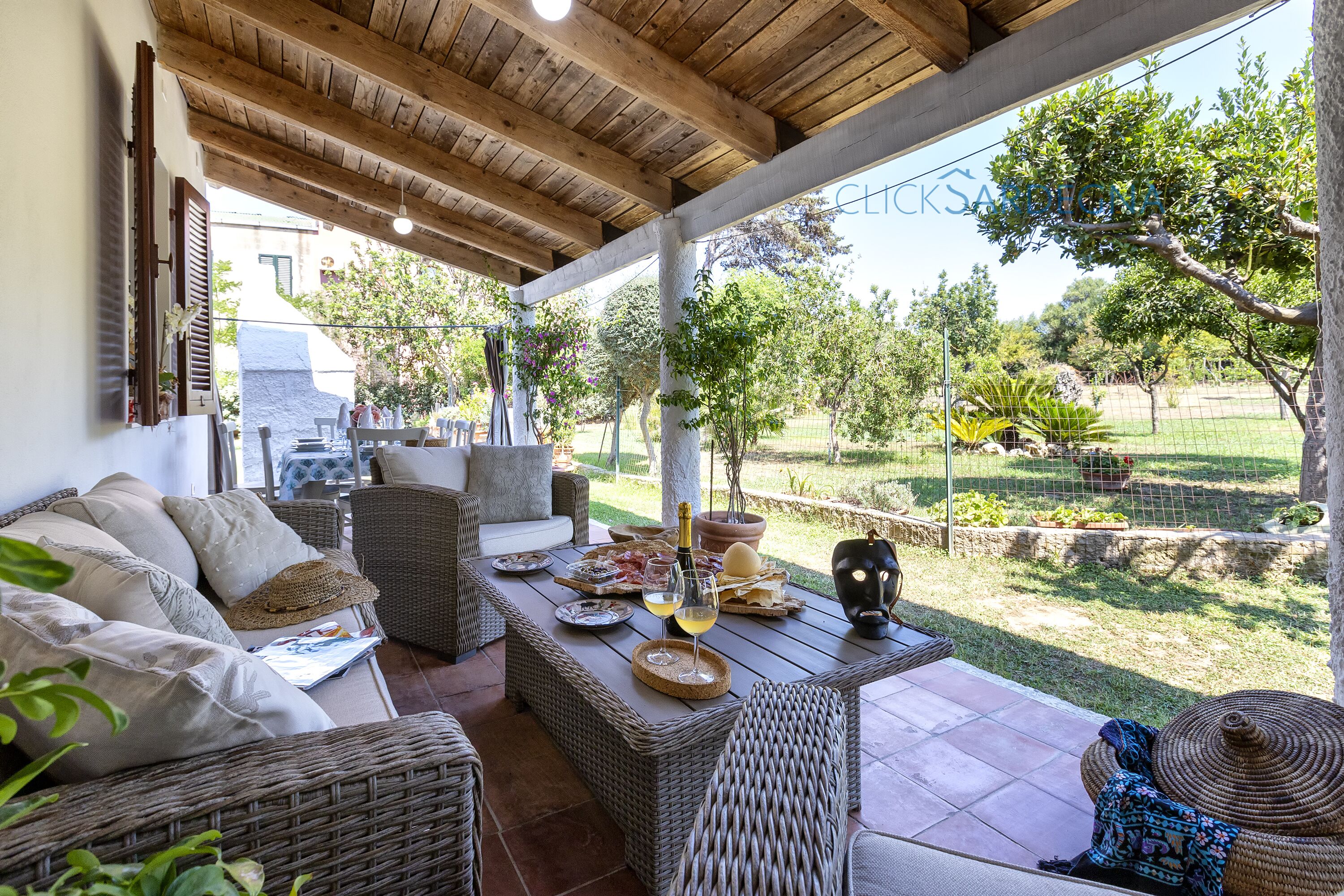 A covered patio with wicker furniture, a table with a bottle and glasses, and a view of the garden.