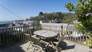A wooden deck with a table and chairs, overlooking a coastal area with houses and a clear sky.