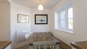 A dining area with a wooden table and white chairs, a framed picture on the wall, and a window with white shutters.