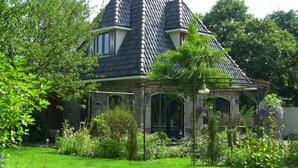 Une maison traditionnelle, dotée d’un toit à forte pente, entourée d’une végétation luxuriante et de plantes en fleurs.