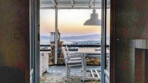 A covered outdoor seating area with stone flooring, a table, and chairs, overlooking a scenic landscape with mountains and a body of water.