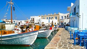 A narrow street lined with boats docked along the water, white buildings with blue accents, and outdoor seating areas.