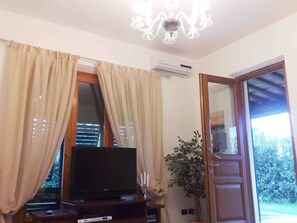 A living room with a flat-screen TV on a wooden stand, beige curtains, a chandelier, and a door leading outside.