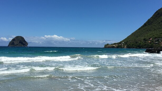 Une plage aux eaux turquoise et limpides, un promontoire rocheux et une colline verdoyante parsemée de bâtiments.