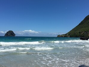 Une plage aux eaux turquoise et limpides, un promontoire rocheux et une colline verdoyante parsemée de bâtiments.