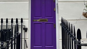 A purple door with a brass knocker, a white wall with a circular sign above, and two black lantern-style lights on either side.