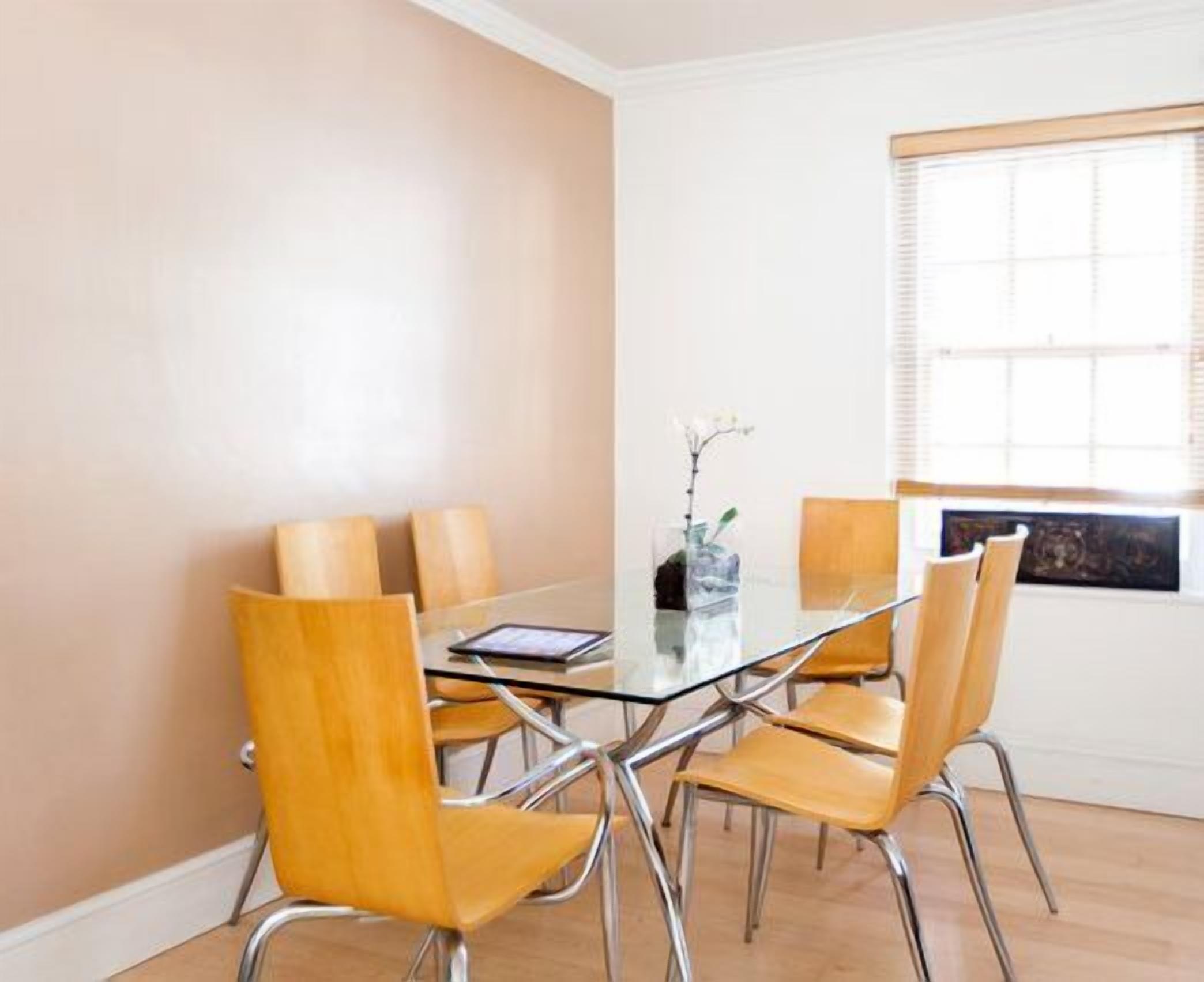 A dining area with a glass table surrounded by four wooden chairs, a vase with flowers, and a window with blinds.