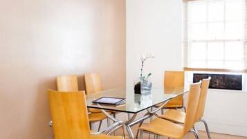 A dining area with a glass table surrounded by four wooden chairs, a vase with flowers, and a window with blinds.