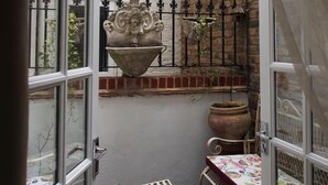 A narrow courtyard with a white brick wall, a metal gate, a large decorative pot, a small table with a colorful tablecloth, and a chair.