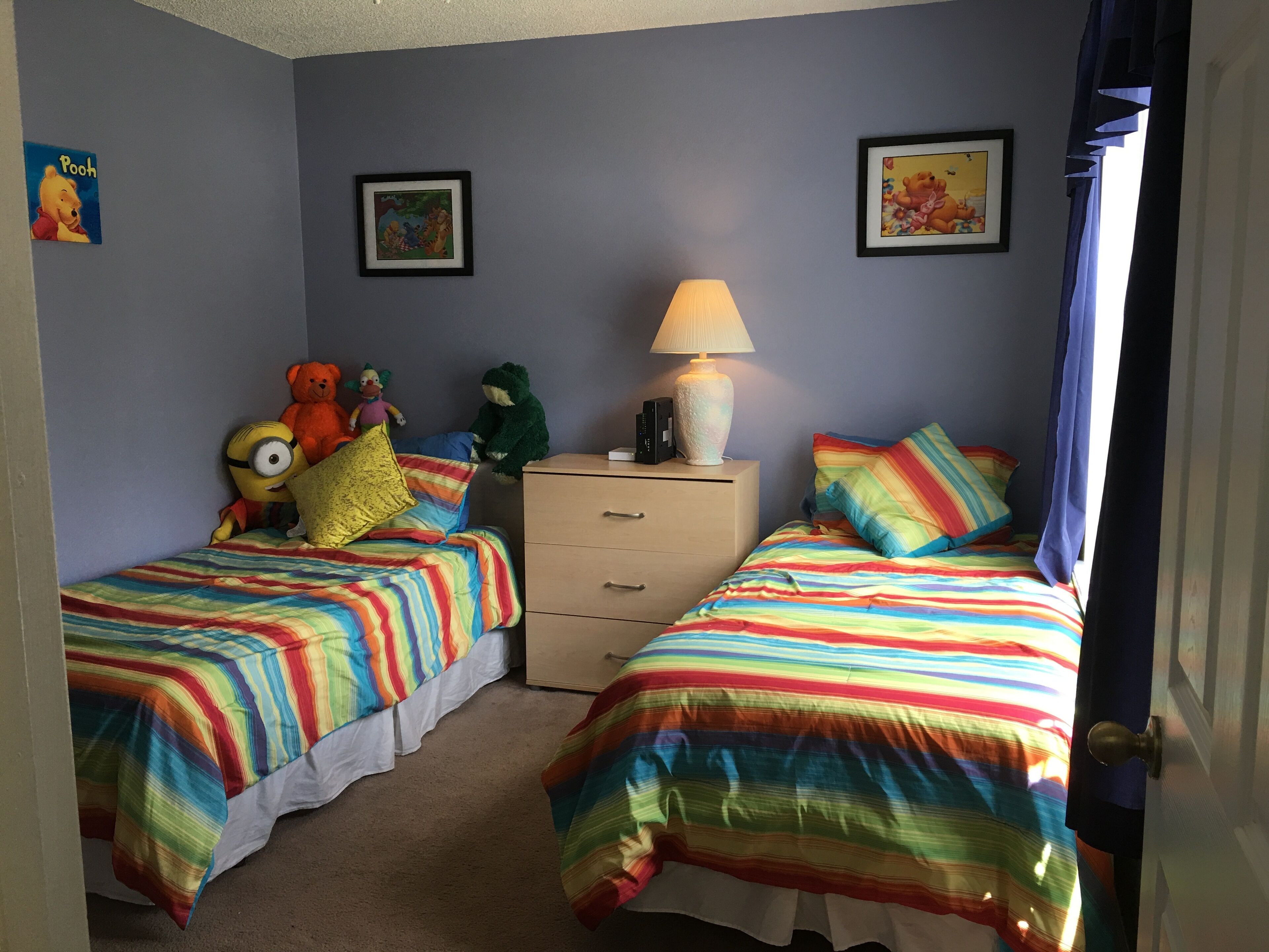 A bedroom with two beds, a nightstand with a lamp, and plush toys on the left side.