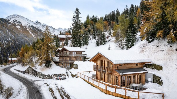 Una cabaña de madera con balcón, rodeada de árboles y montañas cubiertas de nieve.