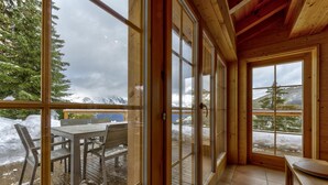 Una cabaña de madera con ventanas amplias que ofrecen vistas a montañas nevadas. En el interior, hay una mesa y sillas de comedor.