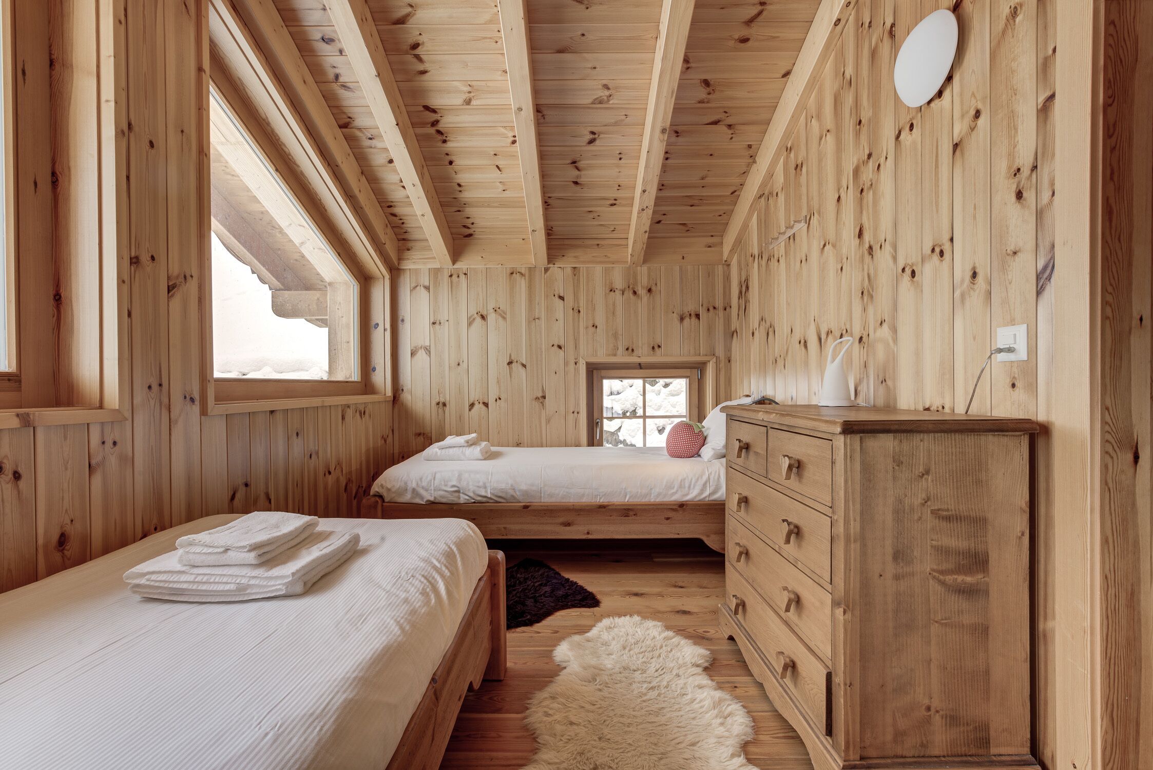 A wooden cabin bedroom with two beds, a dresser, and a large window.