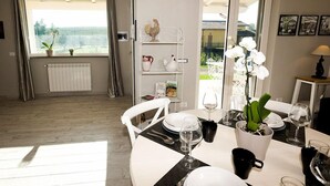A dining area with a table set for two, a radiator, a window with curtains, a shelf with decorative items, and a view of the outdoors.