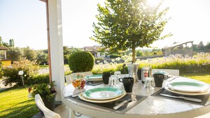 An outdoor dining table set with plates, cutlery, and a coffee pot, surrounded by chairs and a potted plant.