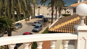 Een kuststraat met geparkeerde auto's, palmbomen en een strand op de achtergrond.