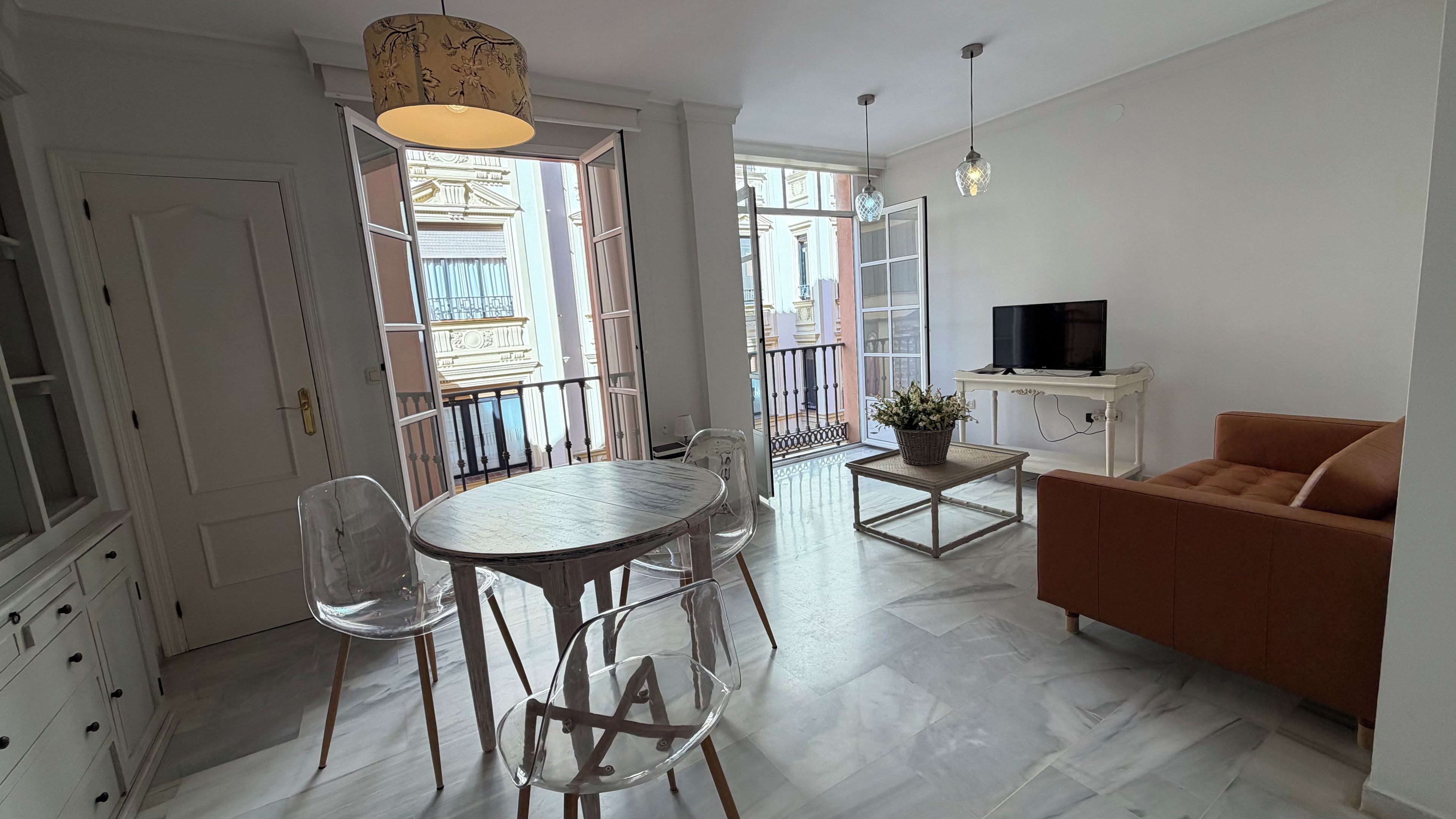 A modern living room with a dining area, a sofa, a TV, and a balcony with a view of buildings.