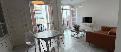 A modern living room with a dining area, a sofa, a TV, and a balcony with a view of buildings.