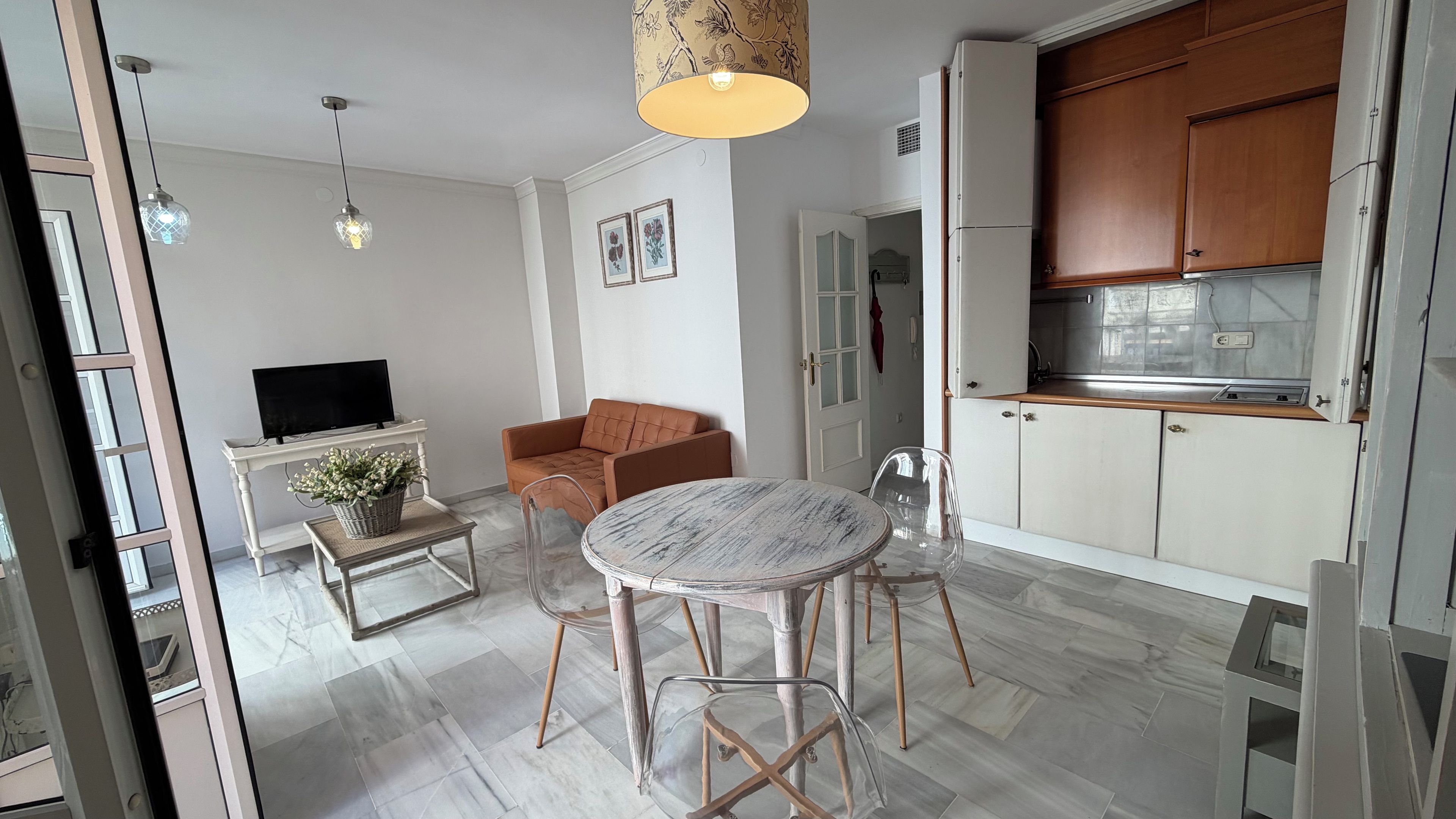 A modern kitchen with wooden cabinets, a dining table with chairs, a sofa, and a television on a stand.