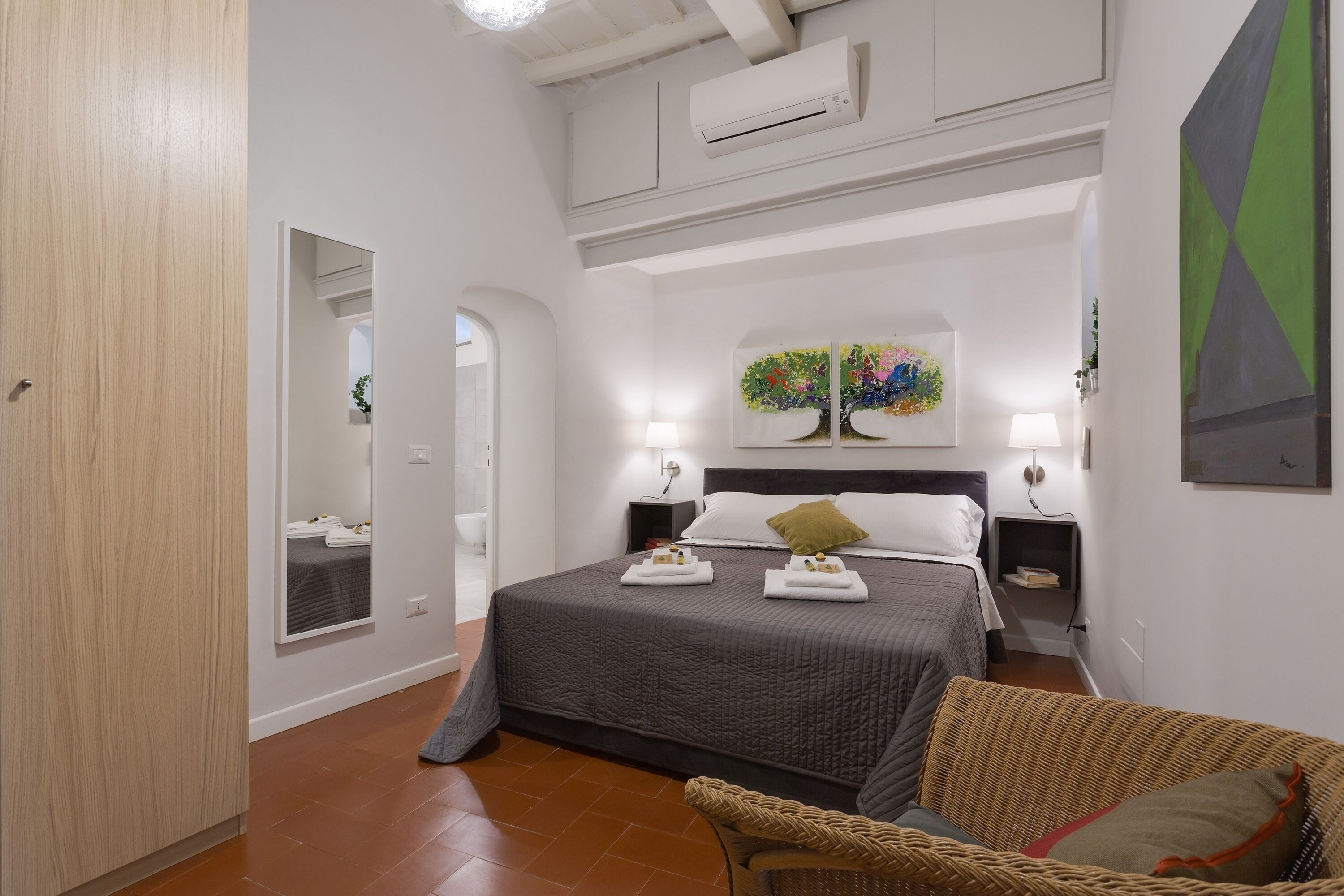A bedroom with a large bed, two bedside tables, a mirror, a wall-mounted air conditioner, and a painting above the bed.