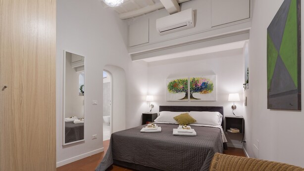 A bedroom with a large bed, two bedside tables, a mirror, a wall-mounted air conditioner, and a painting above the bed. - Grotta Pinta 7 | Cozy and romantic (Roma)