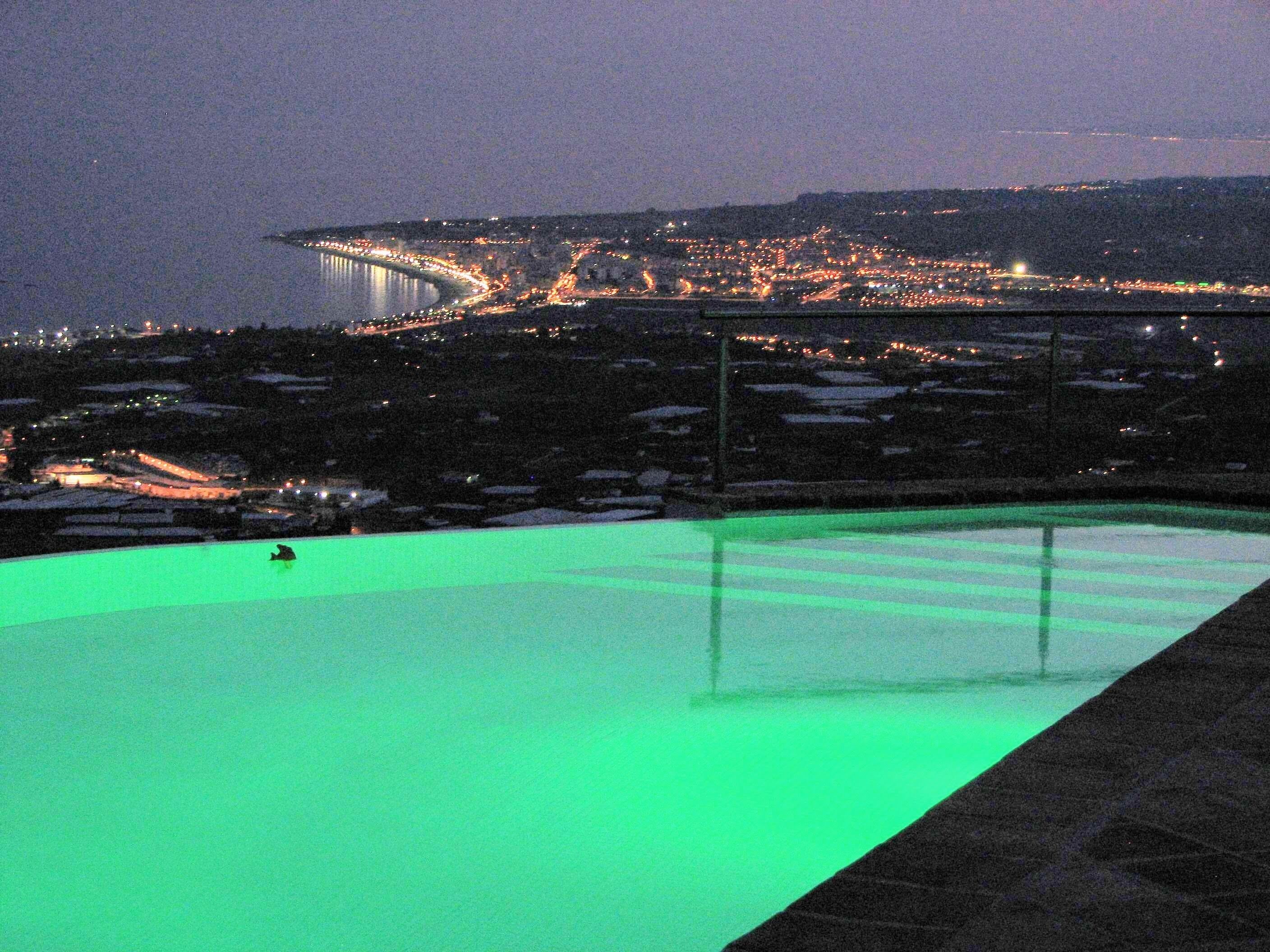 A nighttime view of a pool with a green glow, surrounded by a cityscape with distant lights and a bridge.