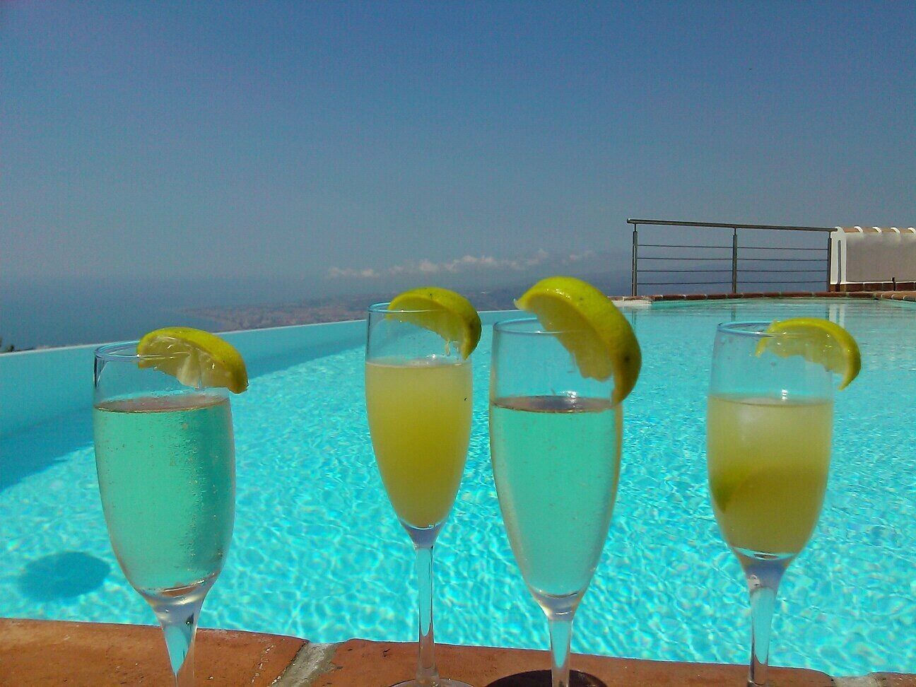 Tre champagneglas med citronskivor står på ett bord vid poolkanten, med fri utsikt över havet.