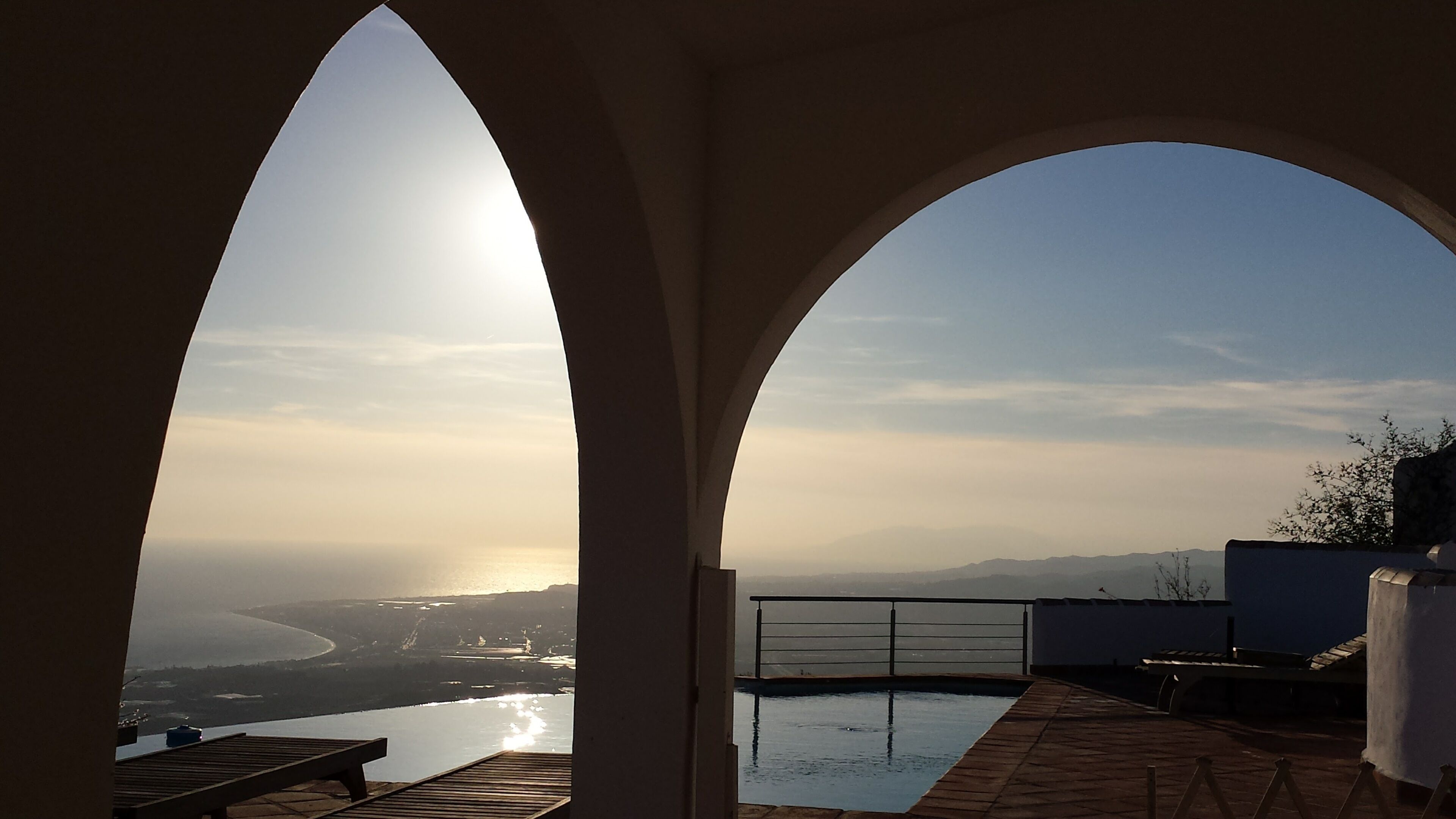 A view through an arched window overlooking a body of water and distant landscape, with a pool and seating area visible.