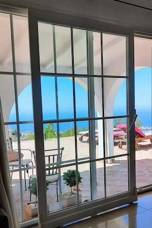 On aperçoit, à travers une porte vitrée, un espace extérieur aménagé en terrasse, avec une piscine, des chaises longues et des plantes en pot.